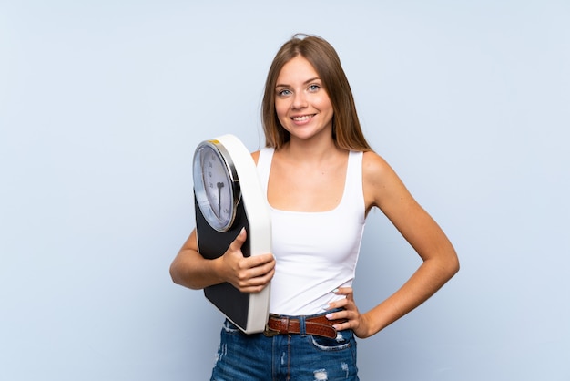 Young blonde girl with weighing machine over isolated blue white wall