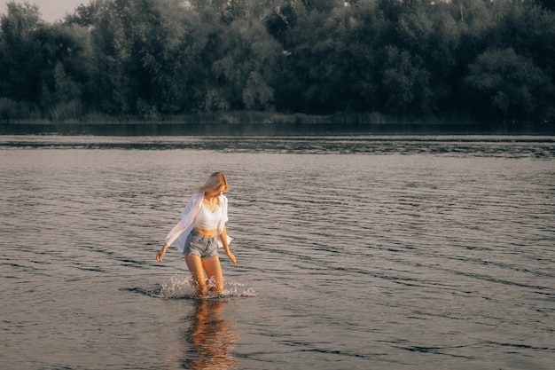 A young blonde is running in the water at dawn. A young woman in a white top, shirt, denim shorts looks at the water and runs along the river against the background of a beautiful landscape.