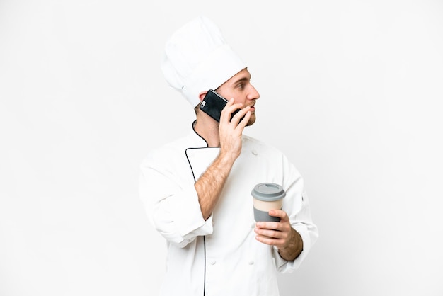 Young blonde man Chef over isolated white background holding coffee to take away and a mobile