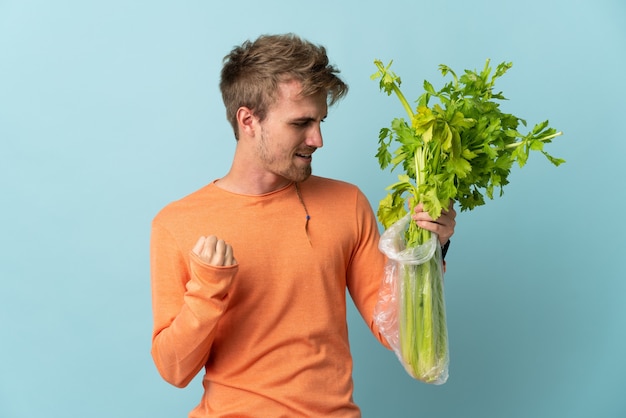 Young blonde man holding a celery isolated on blue background celebrating a victory