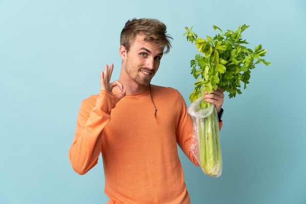 Young blonde man holding a celery isolated on blue background showing ok sign with fingers