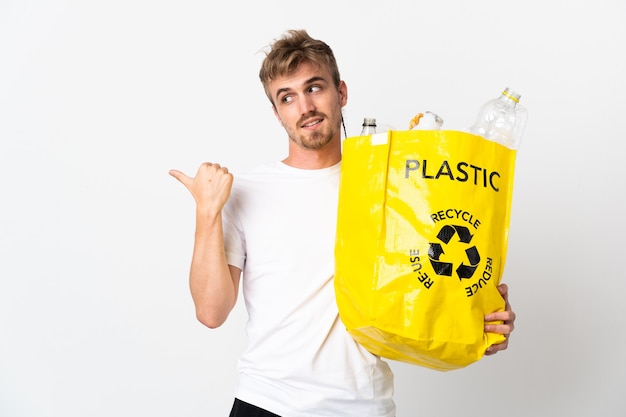 Young blonde man holding a recycling bag full of paper to recycle isolated on white background pointing to the side to present a product