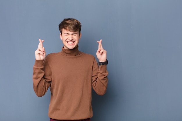 Young blonde man smiling and anxiously crossing both fingers, feeling worried and wishing or hoping for good luck isolated against flat wall