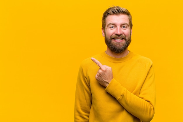 Young blonde man smiling cheerfully, feeling happy and pointing to the side and upwards, showing object in copy space against orange wall