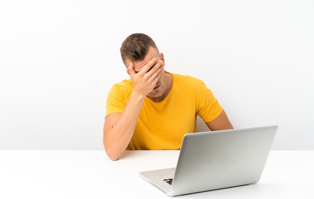 Young blonde man in a table with a laptop covering eyes by hands