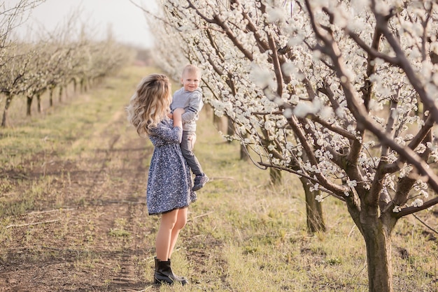 Young blonde mother with her little son walk by the hand among the blossoming apple orchards