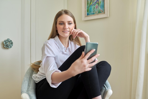 Young blonde sitting at home in chair using video communication on smartphone