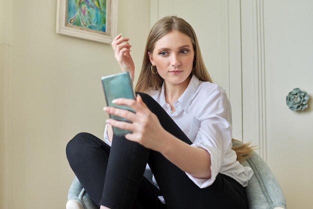 Young blonde sitting at home in chair using video communication on smartphone