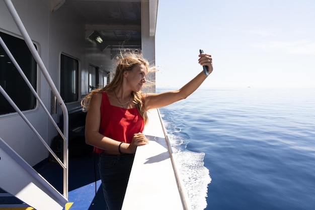 Young blonde tourist taking photos with her smartphone on a ferryboat