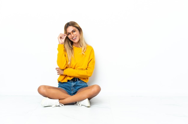 Photo young blonde uruguayan woman sitting on the floor isolated on white wall with glasses and happy