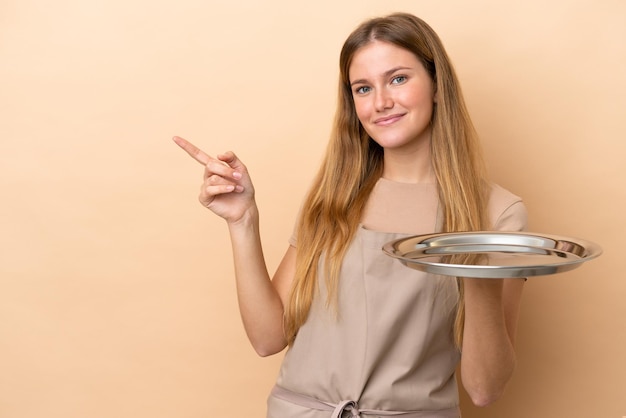 Young blonde waitress woman with tray isolated on beige background pointing finger to the side