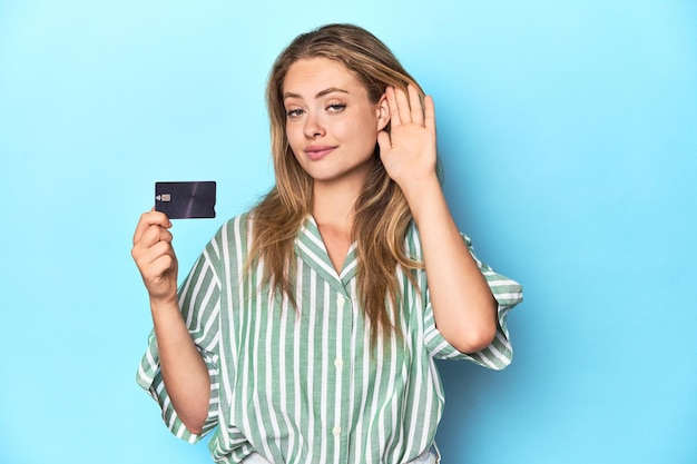 Young blonde with a credit card in blue studio trying to listening a gossip