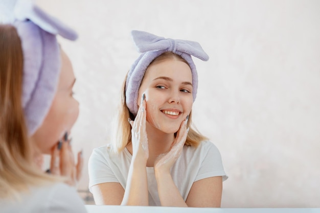 Young blonde woman applies facial cleaner for skin wash teen girl washing face morning in bathroom