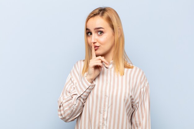 Young blonde woman asking for silence and quiet, gesturing with finger in front of mouth, saying shh or keeping a secret