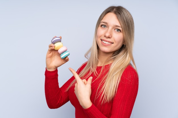 Young blonde woman over blue holding colorful French macarons and pointing it