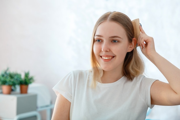 Young blonde woman combing her tangle hair with wooden comb hairbrush.Morning routine in bathroom