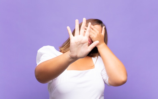 Young blonde woman covering face with hand and putting other hand up front, stop gesture