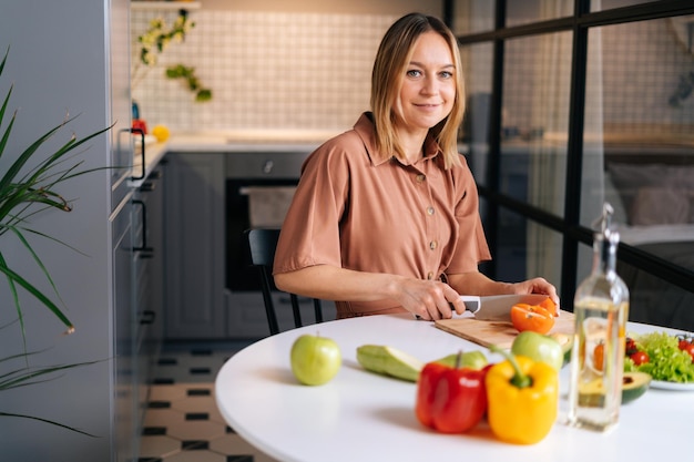 Young blonde woman cutting onion in kitchen with modern interior looking at camera Concept of healthy eating