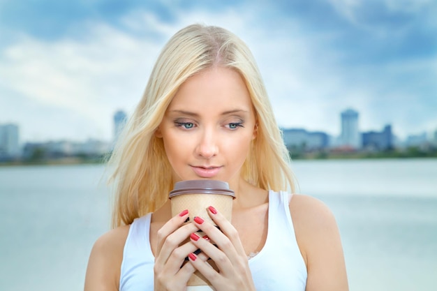 Young blonde woman drinking coffee on a summer morning by the river with views of the city Lifestyle