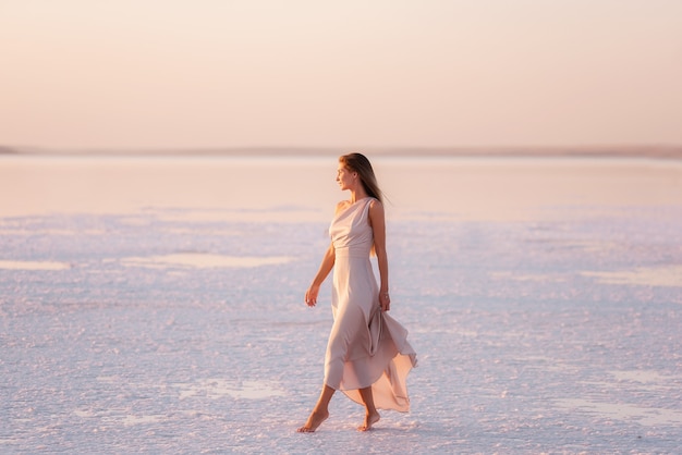 Young blonde woman in an evening airy pastel pink, powdery dress stands barefoot on white crystallized salt.   Salt mining trip, walking on water at sunset