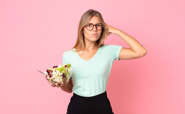 Young blonde woman feeling confused and puzzled, showing you are insane and holding a salad