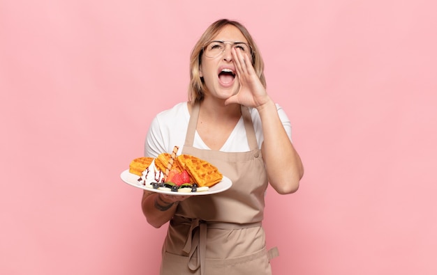 Young blonde woman feeling happy, excited and positive, giving a big shout out with hands next to mouth, calling out