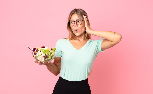 young blonde woman feeling happy, excited and surprised and holding a salad