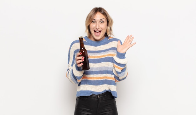 Young blonde woman feeling happy, excited, surprised or shocked, holding a beer