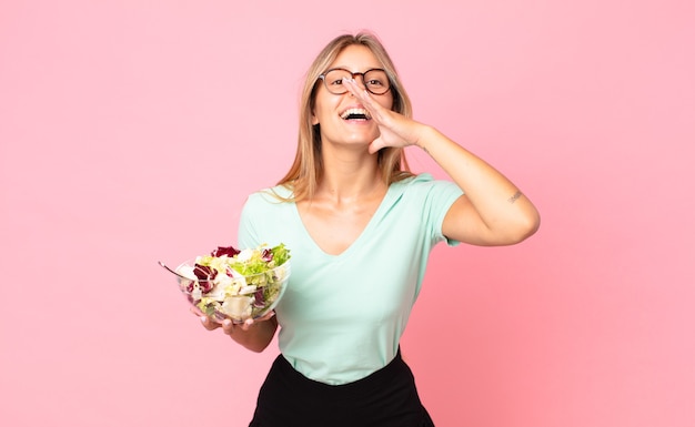 Young blonde woman feeling happy,giving a big shout out with hands next to mouth and holding a salad