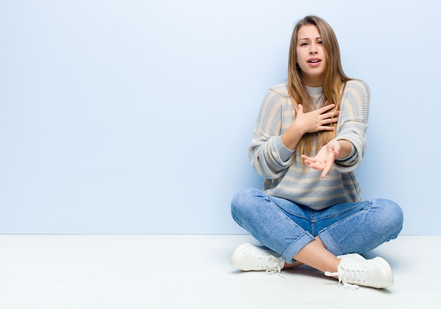 Young blonde woman feeling happy and in love, smiling with one hand next to heart and the other stretched up front sitting on the floor