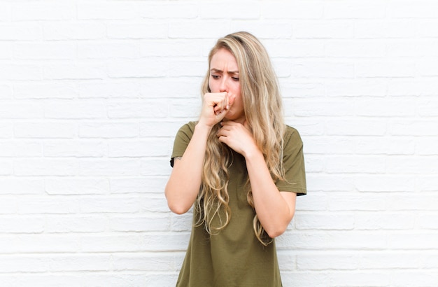 Young blonde woman feeling ill with a sore throat and flu symptoms, coughing with mouth covered against brick wall