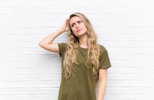 Young blonde woman feeling puzzled and confused, scratching head and looking to the side against brick wall background