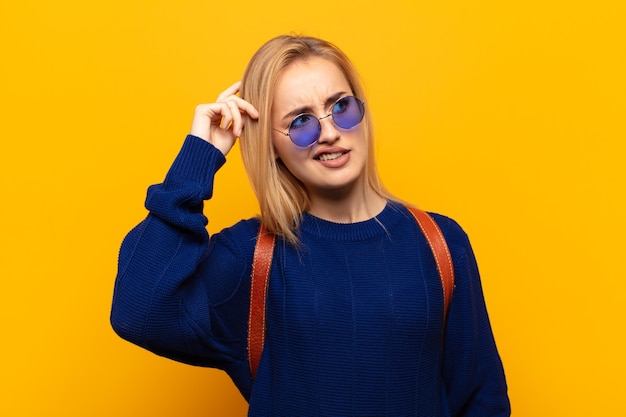 Young blonde woman feeling puzzled and confused, scratching head and looking to the side