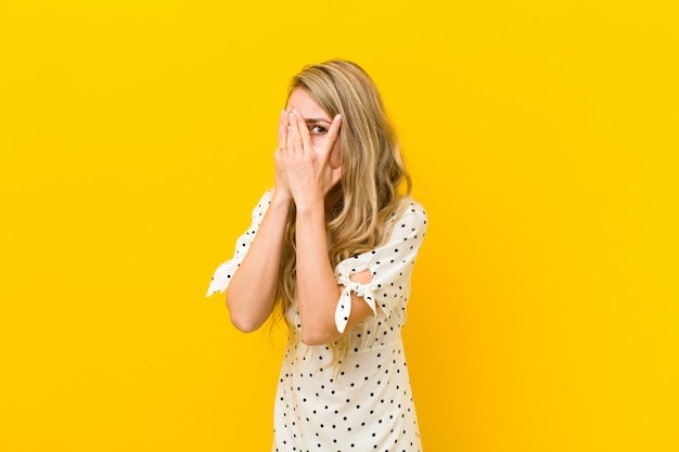 Young blonde woman feeling scared or embarrassed, peeking or spying with eyes half-covered with hands against yellow wall