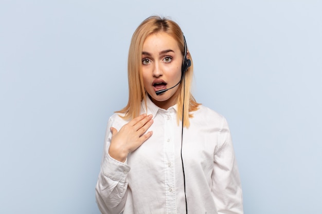 Young blonde woman feeling shocked, astonished and surprised, with hand on chest and open mouth, saying who, me?