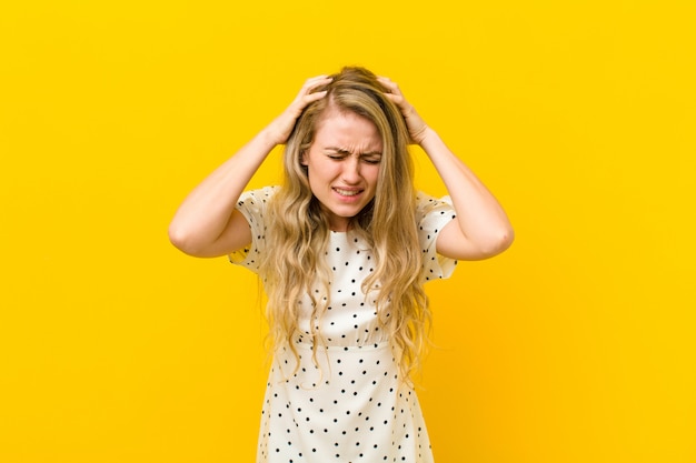 Young blonde woman feeling stressed and frustrated, raising hands to head, feeling tired, unhappy and with migraine over yellow wall