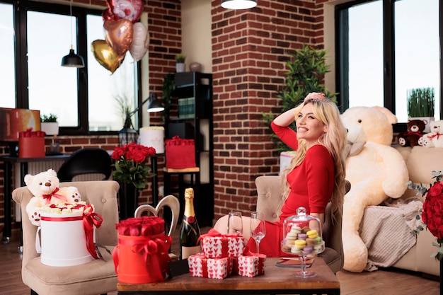 Young blonde woman getting luxury valentines day gifts, sitting in living room filled with expensive present boxes, bear soft toy, red roses, balloons. February 14 girlfriend romantic greeting