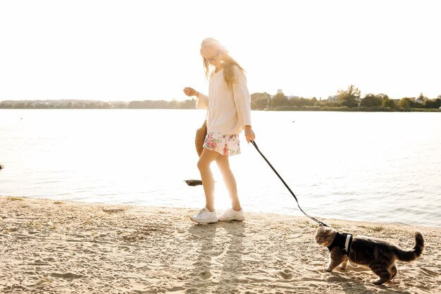 Young blonde woman girl walking on the beach in spring in a dress and white sweater with a Scottish straight cat on a sunny day