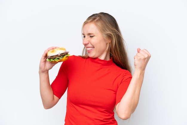 Young blonde woman holding a burger isolated on white background celebrating a victory