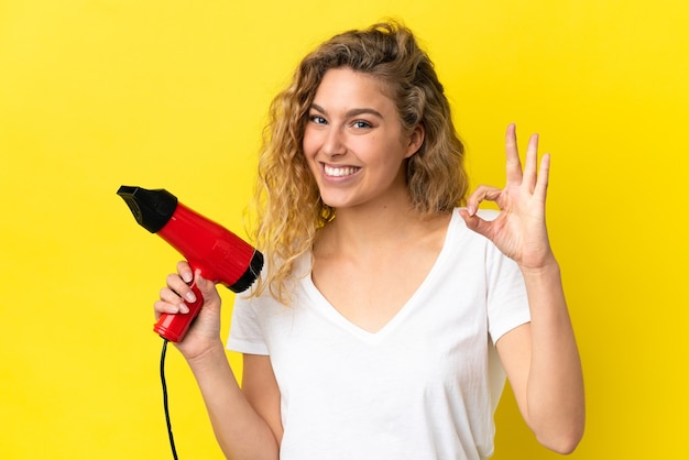 Young blonde woman holding a hairdryer isolated on yellow background showing ok sign with fingers