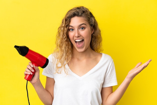 Young blonde woman holding a hairdryer isolated on yellow background with shocked facial expression