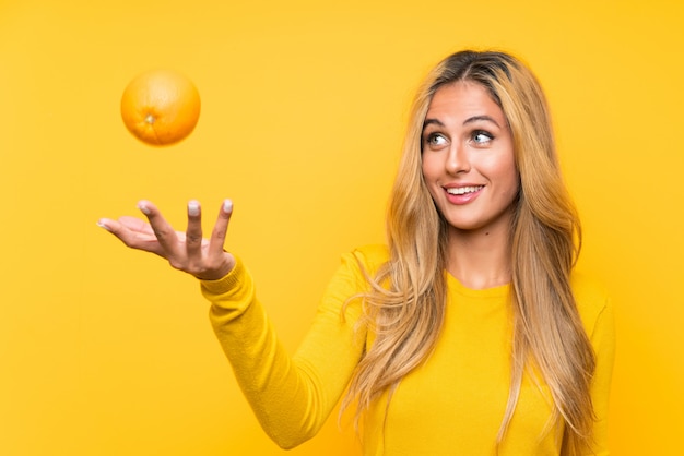 Young blonde woman holding an orange over yellow wall