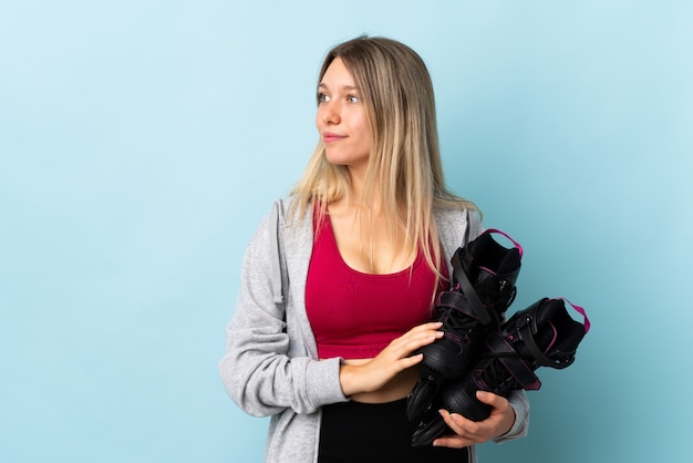 Young blonde woman holding a roller skates isolated on pink wall looking to the side