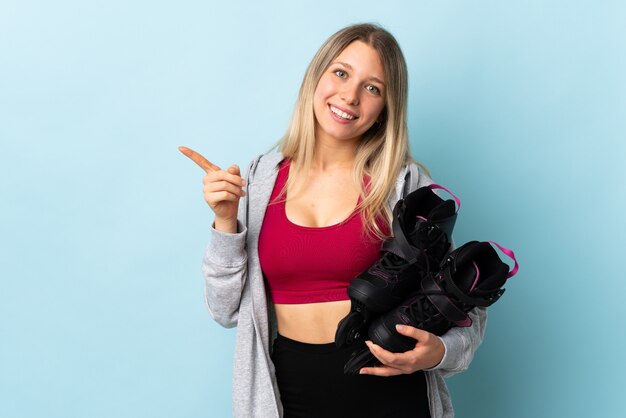 Young blonde woman holding a roller skates isolated on pink wall pointing finger to the side