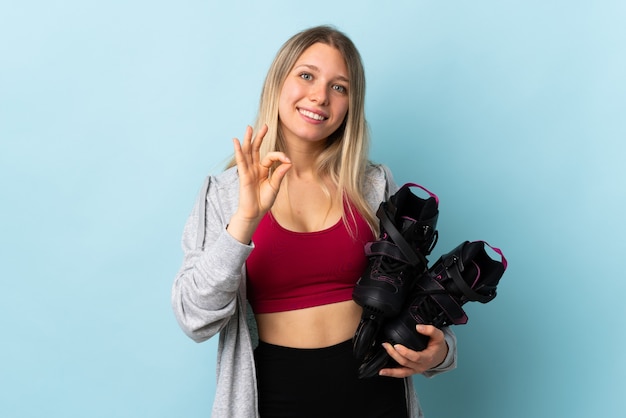 Young blonde woman holding a roller skates isolated on pink wall showing an ok sign with fingers