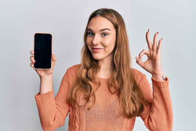 Young blonde woman holding smartphone showing screen doing ok sign with fingers, smiling friendly gesturing excellent symbol