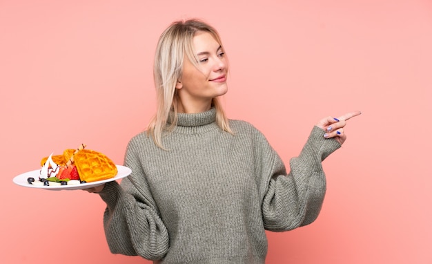 Photo young blonde woman holding waffles over isolated pink wall pointing to the side to present a product