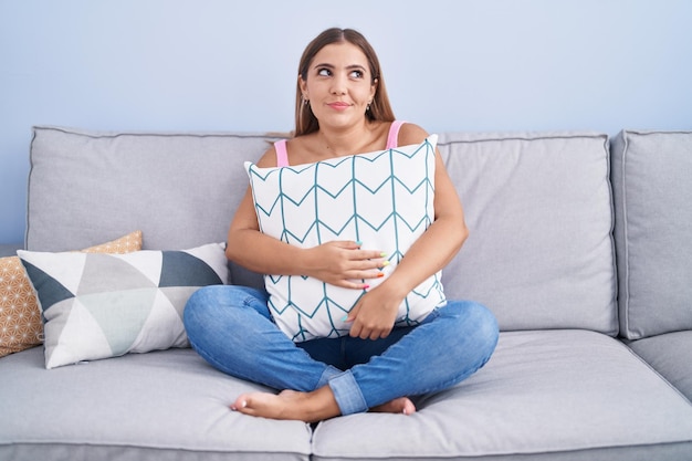 Young blonde woman hugging pillow sitting on the sofa smiling looking to the side and staring away thinking