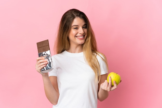 Young blonde woman over isolated background taking a chocolate tablet in one hand and an apple in the other