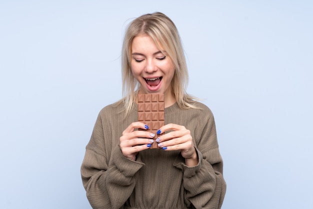 Young blonde woman over isolated blue wall eating a chocolate tablet